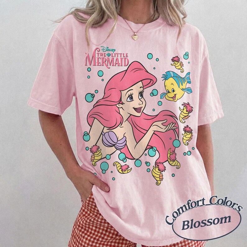 May include: A light pink short-sleeve t-shirt featuring a graphic print of Ariel from The Little Mermaid with long pink hair, a purple seashell top, and the character Flounder. The design includes teal bubbles and small yellow seahorses.