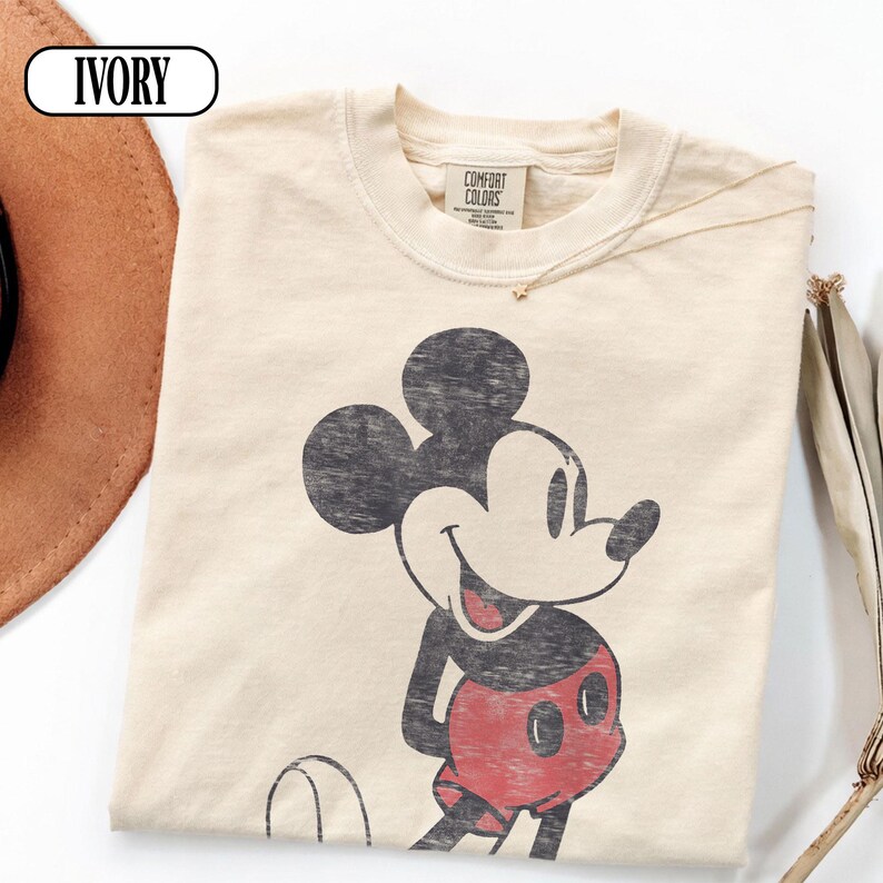 以下が含まれることがあります： ミッキーマウスのヴィンテージグラフィックが特徴のアイボリー色のTシャツ。 シャツはクルーネックと半袖です。 タグには「Comfort Colors」と書かれています。 ゴールドのネックレスが見えます。 「IVORY」という単語が白い楕円の中にあります。