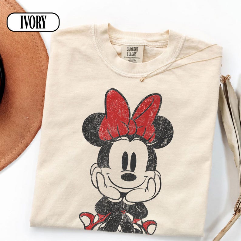 May include: Ivory-colored t-shirt featuring a distressed graphic of Minnie Mouse with a red bow. The shirt has a crew neck and short sleeves. The tag reads "Comfort Colors". A gold necklace is visible. The word "IVORY" is in a white oval in the upper left corner.