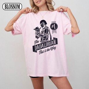 Puede incluir: Camiseta rosa claro con un gr&aacute;fico negro de The Mandalorian sosteniendo a Grogu, con el texto "The DADALORIAN This is the Way". Tambi&eacute;n tiene un sello "#1 in the Galaxy". La marca "BLOSSOM" est&aacute; en la esquina superior izquierda.