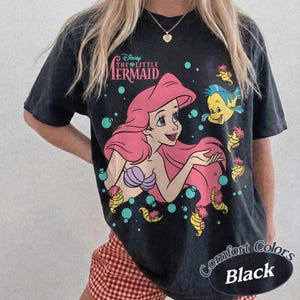 May include: A black short-sleeve t-shirt featuring a graphic of Ariel from The Little Mermaid with long pink hair, a purple seashell top, and Flounder. The text reads Disney The Little Mermaid. Teal bubbles and small yellow sea creatures surround them.
