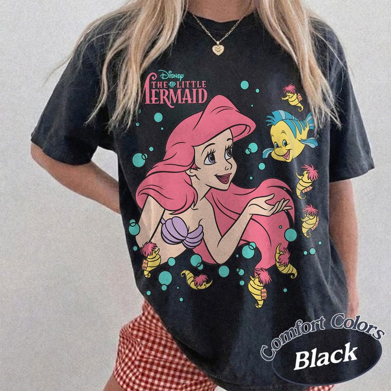 May include: A black short-sleeve t-shirt featuring a graphic of Ariel from The Little Mermaid with long pink hair, a purple seashell top, and Flounder. The text reads Disney The Little Mermaid. Teal bubbles and small yellow sea creatures surround them.