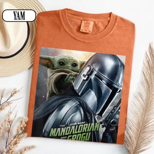 Puede incluir: Camiseta naranja con un gr&aacute;fico de The Mandalorian y Grogu de Star Wars. El dise&ntilde;o incluye el texto "The Mandalorian and Grogu". La camiseta tiene cuello redondo y mangas cortas. Un sombrero de paja es visible en el fondo.