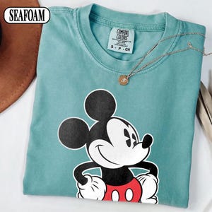 May include: A seafoam green short-sleeve t-shirt featuring a graphic of Mickey Mouse with black ears, a white face, and red shorts. The inner neck label reads Comfort Colors, size S, 100% cotton, made in Honduras. A thin gold chain necklace rests on top.