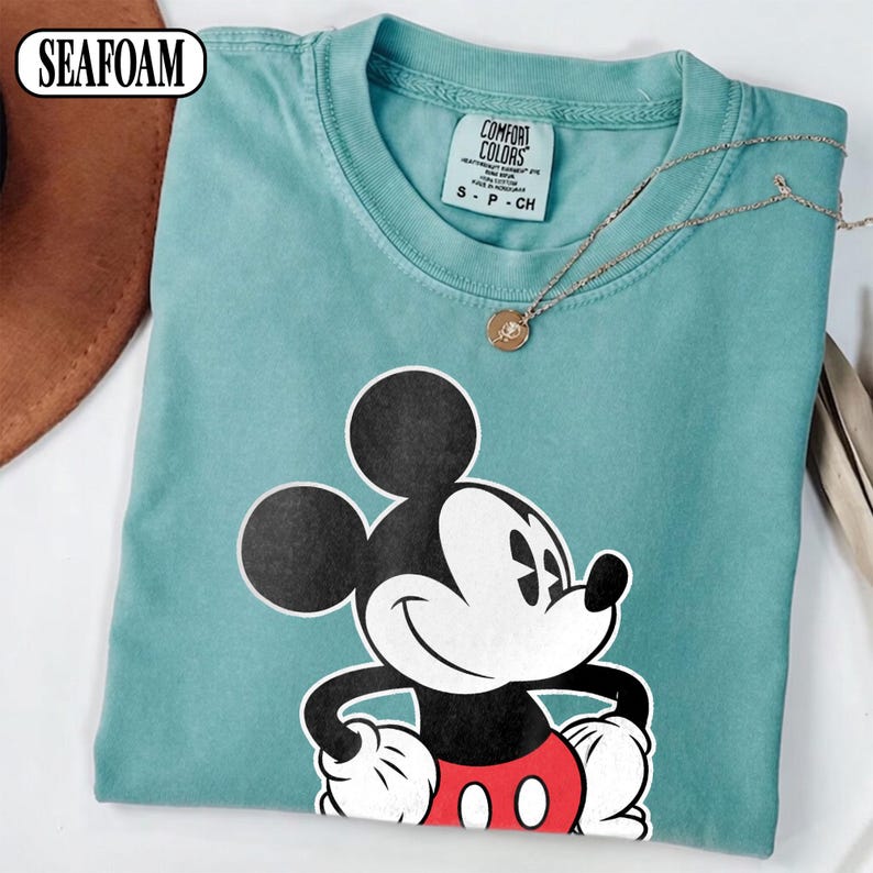 May include: A seafoam green short-sleeve t-shirt featuring a graphic of Mickey Mouse with black ears, a white face, and red shorts. The inner neck label reads Comfort Colors, size S, 100% cotton, made in Honduras. A thin gold chain necklace rests on top.