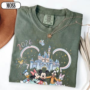 May include: A moss green t-shirt with a Disney-themed graphic, including characters, a castle, and the year 2026. The shirt has a crew neck and short sleeves. A gold necklace with a pendant is draped on the shirt.