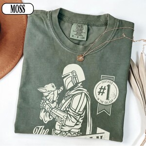 Puede incluir: Camiseta verde musgo con un gr&aacute;fico de The Mandalorian sosteniendo a Grogu, con el texto "Certified #1 in the Galaxy". La camiseta tiene cuello redondo y mangas cortas. Un collar dorado est&aacute; sobre la camiseta.