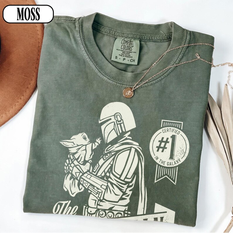 Puede incluir: Camiseta verde musgo con un gr&aacute;fico de The Mandalorian sosteniendo a Grogu, con el texto "Certified #1 in the Galaxy". La camiseta tiene cuello redondo y mangas cortas. Un collar dorado est&aacute; sobre la camiseta.