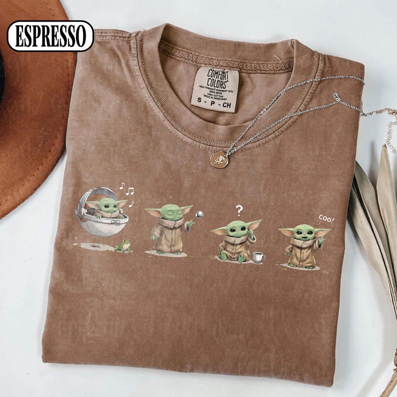 May include: Espresso-colored t-shirt featuring a graphic of a character in various poses. The design includes the character in a pod, holding a ball, with a question mark, and saying "Coo!". The shirt has a Comfort Colors tag.