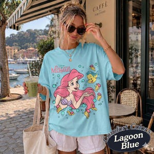 May include: A light blue t-shirt featuring a graphic print of Ariel from The Little Mermaid with pink hair, a purple seashell top, and the yellow fish Flounder. The design includes small bubbles and the text The Little Mermaid in pink lettering.