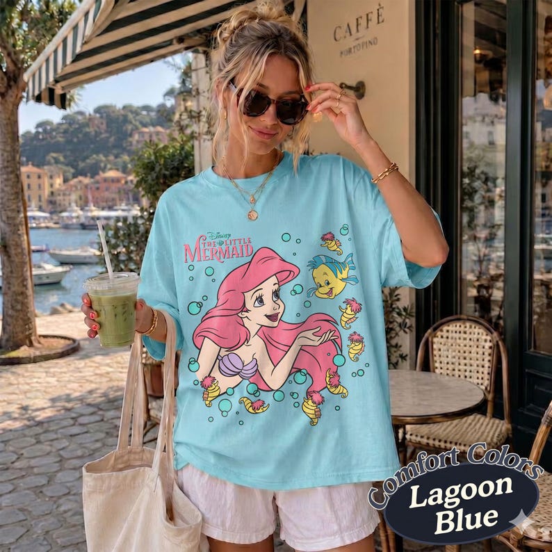 May include: A light blue t-shirt featuring a graphic print of Ariel from The Little Mermaid with pink hair, a purple seashell top, and the yellow fish Flounder. The design includes small bubbles and the text The Little Mermaid in pink lettering.