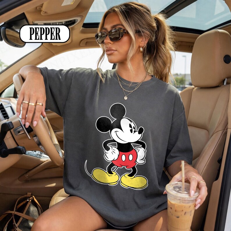 May include: A charcoal gray crewneck t-shirt featuring a large graphic print of Mickey Mouse on the front. The character is depicted in classic colors with black ears, a white face, red shorts, and yellow shoes. The text PEPPER is visible in a box.
