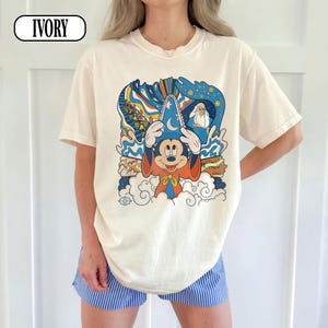 May include: An ivory short-sleeve t-shirt featuring a graphic print of a cartoon mouse wearing a blue wizard hat with a moon and star design, an orange robe, and white gloves, surrounded by blue, orange, and white abstract shapes and clouds.