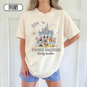 May include: Ivory t-shirt with a Disney-themed graphic, including characters, a castle, and the text "2026 FAMILY VACATION." The top has a crew neck and short sleeves. The fit is relaxed.