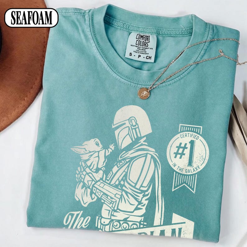 Puede incluir: Camiseta verde mar con un gr&aacute;fico de The Mandalorian sosteniendo a Grogu, con el texto "Certified #1 in the Galaxy". La camiseta tiene cuello redondo y mangas cortas. Un collar plateado con un colgante est&aacute; sobre la camiseta.