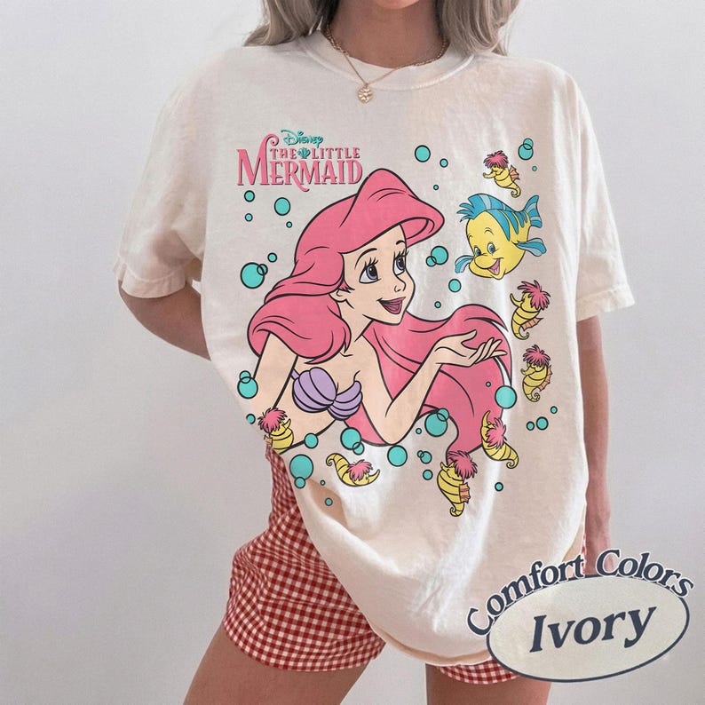 May include: An ivory short-sleeve t-shirt featuring a graphic of Ariel from The Little Mermaid with long pink hair, a purple seashell top, and a blue and yellow fish. The design includes small seahorses and teal bubbles surrounding the characters.