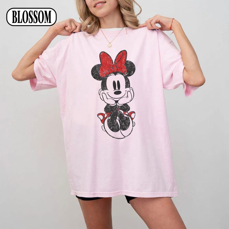 May include: A light pink t-shirt featuring a distressed graphic of Minnie Mouse with a red bow. The shirt has short sleeves and a relaxed fit. The word "BLOSSOM" is in a white oval in the upper left corner.