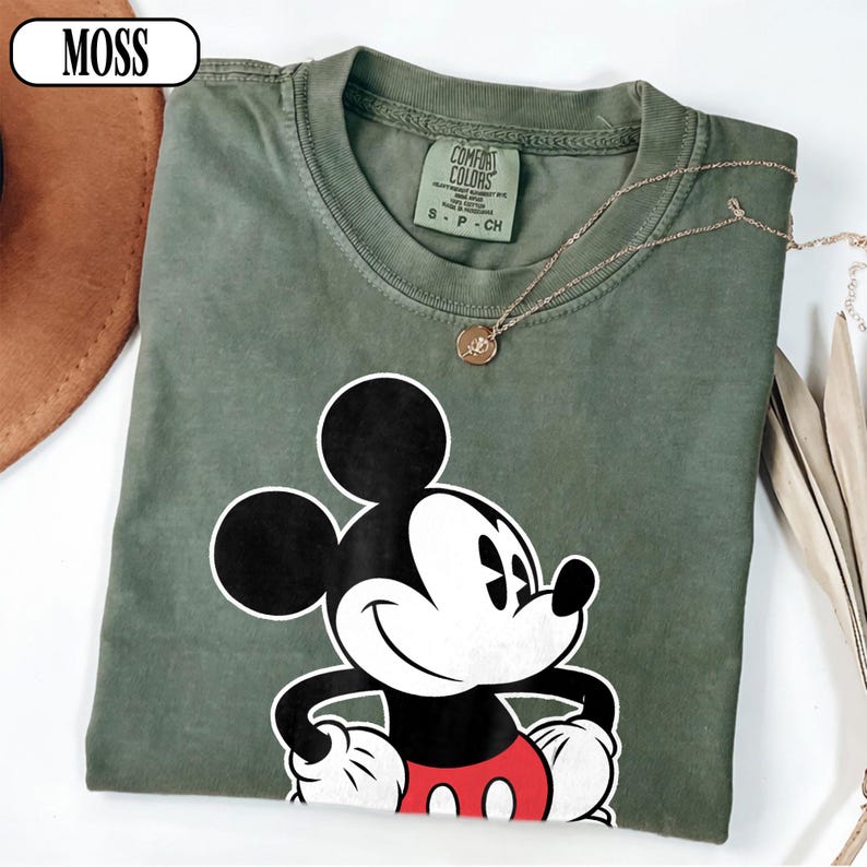 May include: A moss green crew neck t-shirt featuring a graphic of Mickey Mouse with black ears, a white face, and red shorts. The shirt has a Comfort Colors brand label inside the collar and the word MOSS printed in a white box at the top left.