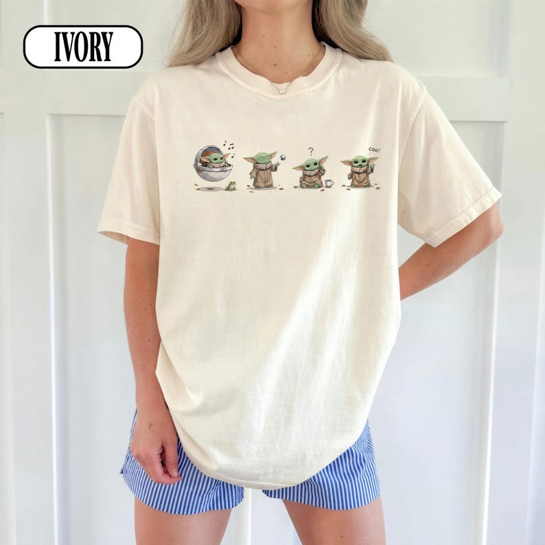 May include: Ivory-colored t-shirt featuring a graphic of Grogu in various poses. The design includes Grogu in a pod, holding a ball, with a question mark, and saying "cool". The shirt has short sleeves and a crew neck.