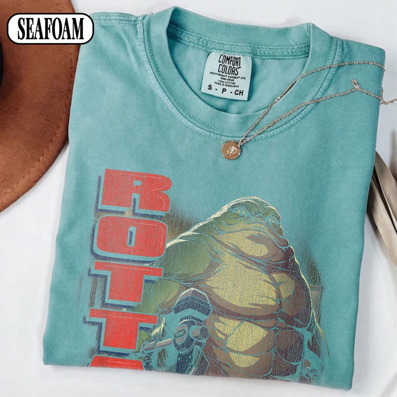 Puede incluir: Camiseta verde espuma de mar con un gr&aacute;fico de una criatura grande y la palabra "ROTTA" en letras rojas. La camiseta tiene una etiqueta Comfort Colors y un peque&ntilde;o collar dorado. La camiseta es talla S-CH.