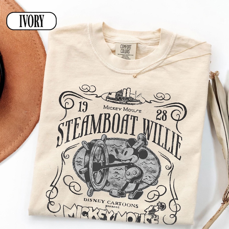 May include: An ivory t-shirt featuring a black graphic print of Steamboat Willie. The design includes the text 1928, Steamboat Willie, Mickey Mouse, and Disney Cartoons present. The shirt has a crew neckline and a Comfort Colors brand label.
