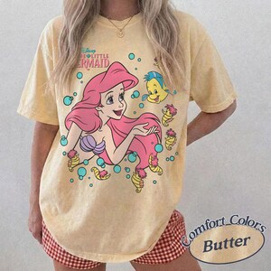 May include: A pale yellow short-sleeve t-shirt featuring a graphic of Ariel from The Little Mermaid with pink hair, a purple seashell top, and Flounder. The design includes blue bubbles and small yellow seahorses surrounding the characters.