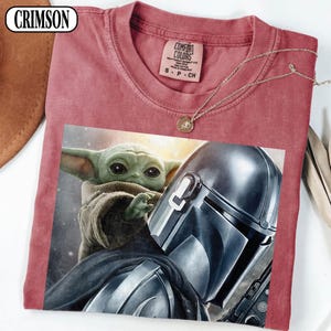 Puede incluir: Camiseta de color carmes&iacute; con un gr&aacute;fico de Grogu y el Mandaloriano. La camiseta tiene cuello redondo y mangas cortas. Un collar de oro con un colgante est&aacute; drapeado sobre la camiseta. La camiseta tiene una etiqueta Comfort Colors.