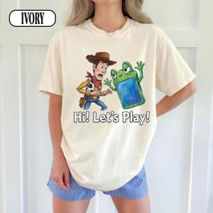 May include: An ivory t-shirt featuring a graphic of a cowboy character in a brown hat and a green frog character holding a blue tablet. The text "Hi! Let's Play!" is printed below the characters. The word "IVORY" appears in a box at the top left.