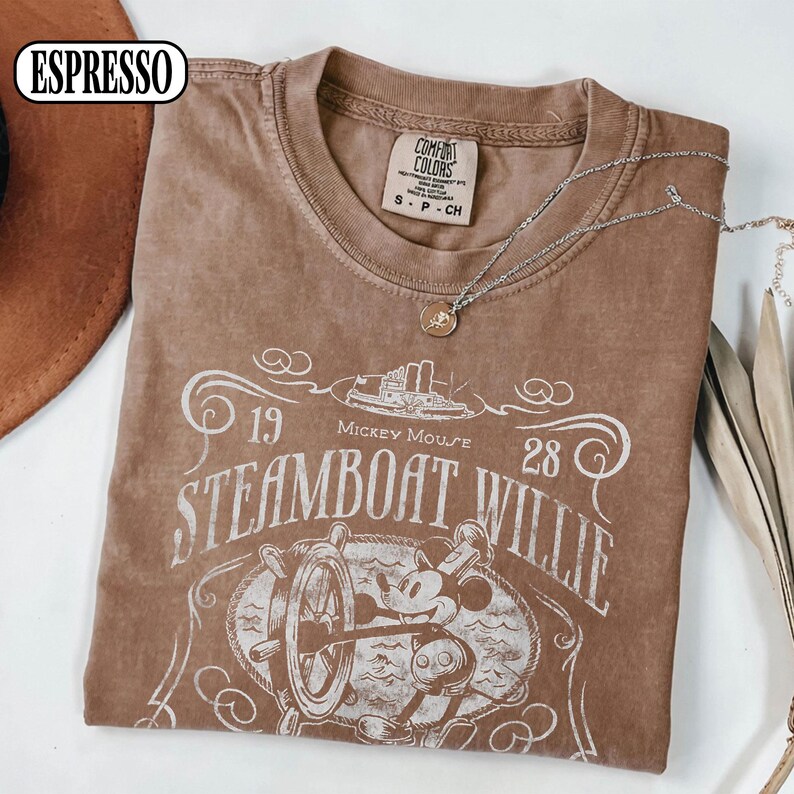 May include: A brown short-sleeve t-shirt featuring a white graphic print of Mickey Mouse at a ship's wheel with the text Steamboat Willie, 1928, and a steamboat illustration. The shirt has a Comfort Colors label in the neck and a size S tag.