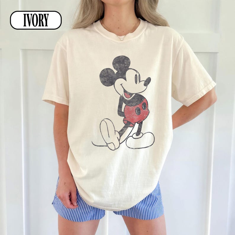 以下が含まれることがあります： アイボリー色のTシャツ。黒、赤、白で描かれたミッキーマウスのヴィンテージ風グラフィックが特徴です。シャツはクラシックなクルーネックと半袖です。「IVORY」の文字が左上に白い楕円でプリントされています。