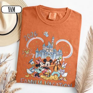 May include: Orange Comfort Colors t-shirt with a Disney-themed graphic. The design includes Mickey Mouse, Minnie Mouse, Donald Duck, Daisy Duck, Goofy, and Pluto, along with a castle and the text "2026 FAMILY VACATION". A straw hat is visible in the background.
