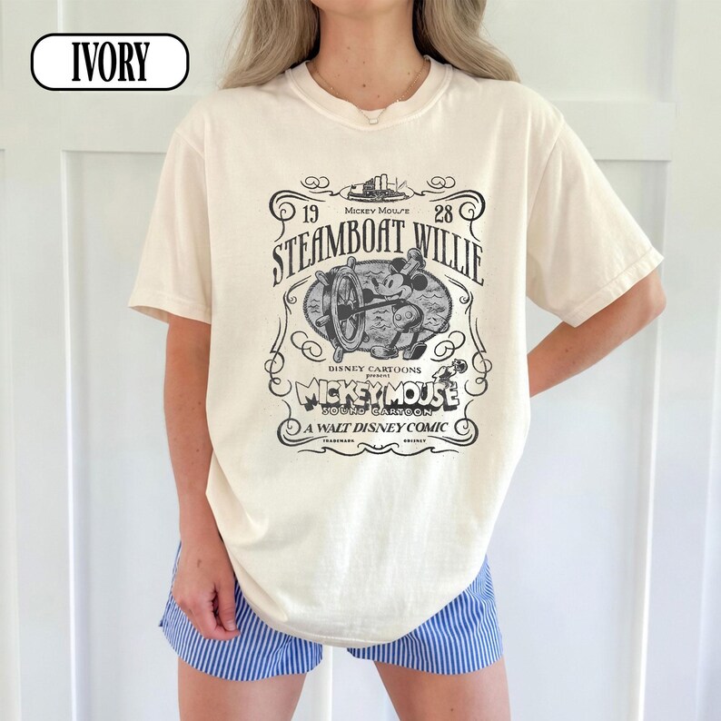 May include: An ivory short-sleeve t-shirt featuring a black graphic print of Steamboat Willie. The design includes the text 1928, Steamboat Willie, Disney Cartoons present, Mickey Mouse sound cartoon, and A Walt Disney Comic with a ship wheel.