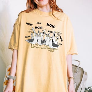 以下が含まれることがあります： マスタードイエローのTシャツ。白と黒の海鳥の漫画イラストが描かれており、足とくちばしは黄色です。「MOM！」の文字が海鳥の周りに繰り返されています。Tシャツは無地です。