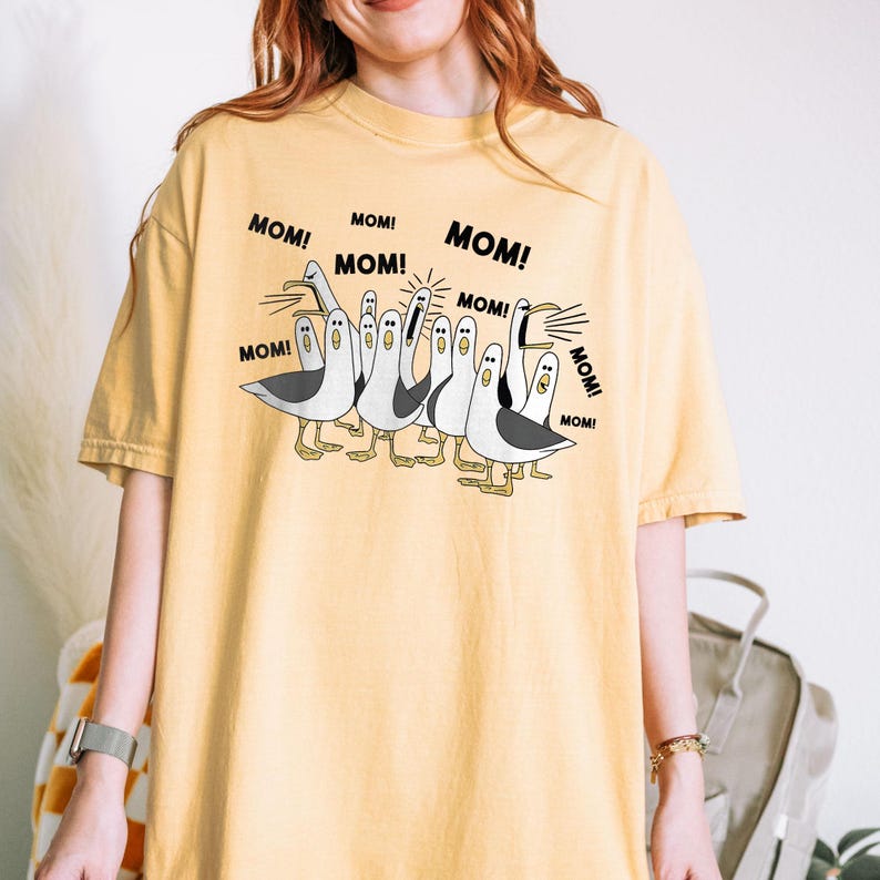 以下が含まれることがあります： マスタードイエローのTシャツ。白と黒の海鳥の漫画イラストが描かれており、足とくちばしは黄色です。「MOM！」の文字が海鳥の周りに繰り返されています。Tシャツは無地です。