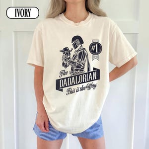 Puede incluir: Camiseta de color marfil con un gr&aacute;fico negro de un personaje sosteniendo una peque&ntilde;a criatura, con el texto "The Dadalorian This is the Way". Una insignia circular dice "#1 in the Galaxy."