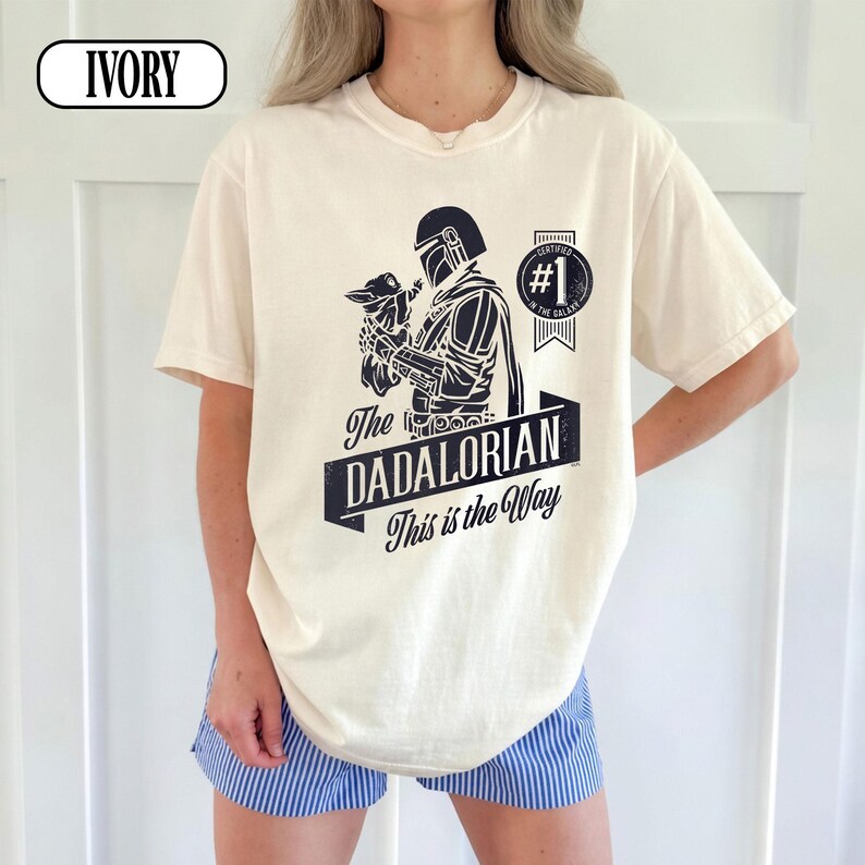 Puede incluir: Camiseta de color marfil con un gr&aacute;fico negro de un personaje sosteniendo una peque&ntilde;a criatura, con el texto "The Dadalorian This is the Way". Una insignia circular dice "#1 in the Galaxy."
