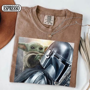 Puede incluir: Camiseta color espresso con un estampado de The Mandalorian y Grogu. La camiseta tiene cuello redondo y mangas cortas. Un collar plateado con un peque&ntilde;o colgante est&aacute; sobre la camiseta. La camiseta es de material suave.