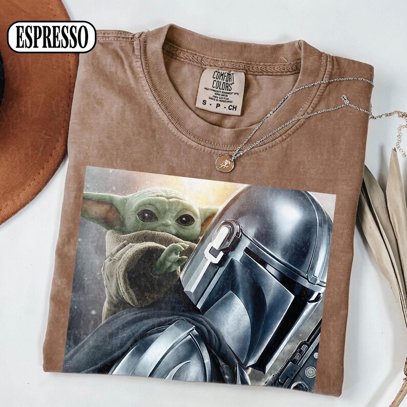 Puede incluir: Camiseta color espresso con un estampado de The Mandalorian y Grogu. La camiseta tiene cuello redondo y mangas cortas. Un collar plateado con un peque&ntilde;o colgante est&aacute; sobre la camiseta. La camiseta es de material suave.
