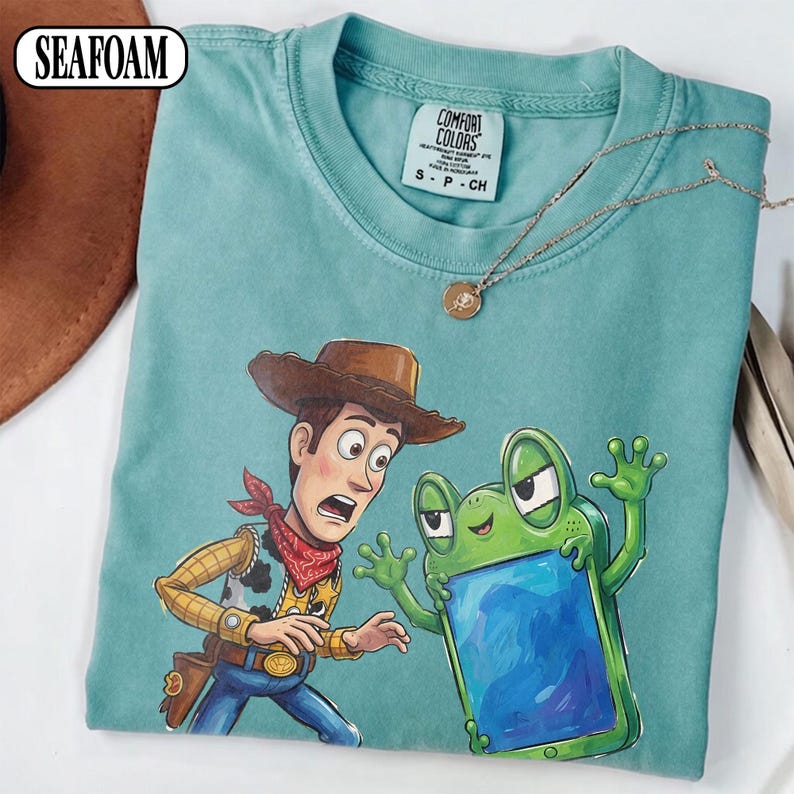 May include: A seafoam green crew neck t-shirt featuring a graphic print of a cowboy character with a brown hat and red bandana looking surprised at a green frog character holding a blue rectangular device. The shirt has a Comfort Colors brand label.