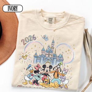May include: Ivory t-shirt with a Disney-themed graphic featuring Mickey Mouse, Minnie Mouse, Donald Duck, Goofy, and Daisy Duck. The design includes a castle, fireworks, a rainbow, and the year "2026". The shirt has a Comfort Colors label.