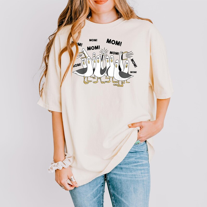 以下が含まれることがあります： クリーム色のTシャツ。漫画の鳥のグラフィックがあり、それぞれに「MOM！」という吹き出しがあります。カモメは白黒で、足は黄色です。ゆったりとしたフィット感のシャツです。