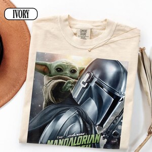 Puede incluir: Camiseta de color marfil con un gr&aacute;fico de The Mandalorian y Grogu de Star Wars. El dise&ntilde;o incluye el casco del Mandaloriano y la cara de Grogu. El texto "THE STAR WARS MANDALORIAN" est&aacute; impreso en la parte inferior en verde y blanco.