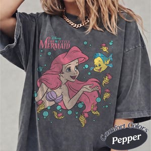 May include: A charcoal gray oversized t-shirt featuring a graphic print of Ariel from The Little Mermaid with long pink hair, a purple seashell top, and Flounder the fish. The design includes teal bubbles and small yellow seahorses.