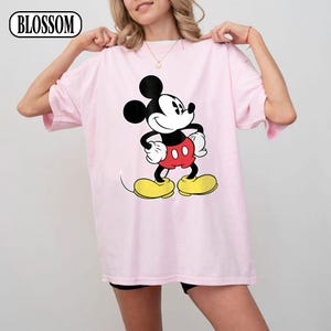 May include: A light pink short-sleeve t-shirt featuring a large graphic of Mickey Mouse in the center. The character is depicted with black ears, a white face, red shorts, and yellow shoes. The word BLOSSOM is printed in the top left corner.