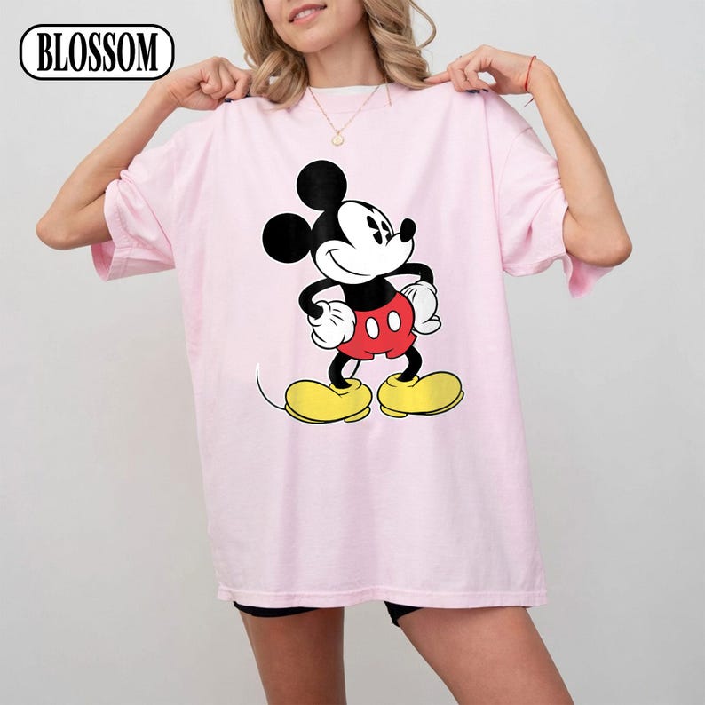 May include: A light pink short-sleeve t-shirt featuring a large graphic of Mickey Mouse in the center. The character is depicted with black ears, a white face, red shorts, and yellow shoes. The word BLOSSOM is printed in the top left corner.