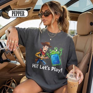 May include: A charcoal grey crewneck t-shirt featuring a graphic print of a cowboy character and a green frog-like creature with a blue screen. The text "Hi! Let's Play!" is printed in white below the characters. The shirt has a relaxed fit.