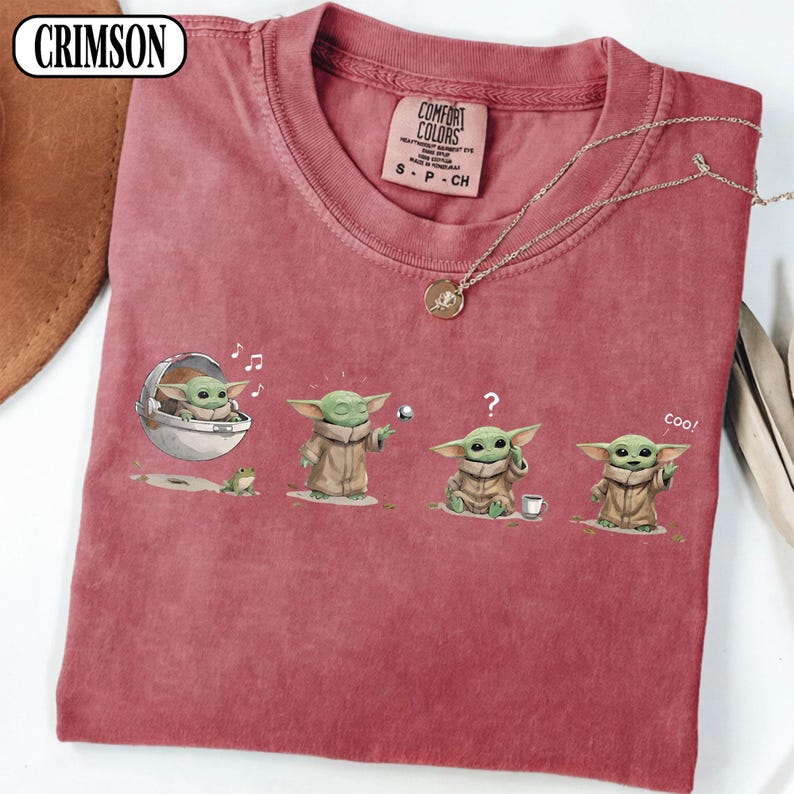 May include: Reddish-pink t-shirt featuring a series of Grogu illustrations. The design includes Grogu in a pod, with a frog, playing with a ball, and drinking from a cup. The shirt has a Comfort Colors tag.