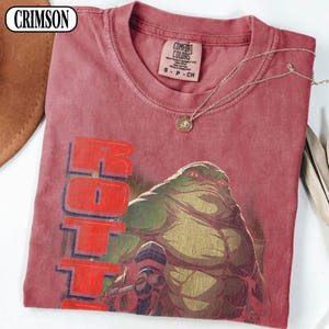 Puede incluir: Camiseta de color rojo carmes&iacute; con un gr&aacute;fico de un alien&iacute;gena verde y marr&oacute;n y la palabra "HOTT" en letras rojas. La camiseta tiene cuello redondo y una etiqueta peque&ntilde;a. Un collar plateado con un colgante est&aacute; drapeado sobre la camiseta.
