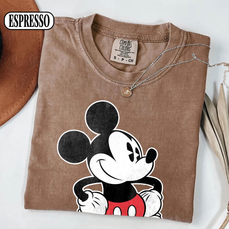 May include: A brown short-sleeved t-shirt featuring a graphic of Mickey Mouse with black ears, a white face, and red shorts. The shirt has a crew neckline and a Comfort Colors brand label inside the collar. The word ESPRESSO is printed in the corner.