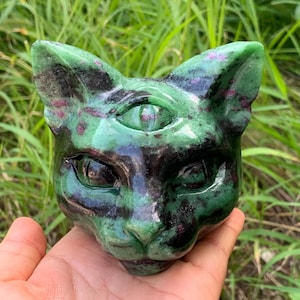 May include: A carved stone cat head sculpture featuring a third eye on the forehead. The object is primarily green with dark black patches and small magenta-colored inclusions throughout the polished surface, showcasing a feline facial structure.